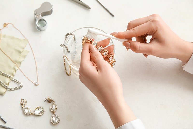 Woman cleaning beautiful bracelet with toothbrush on light background,